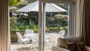 Un patio con una mesa y sillas, un ventanal corredero que da a un jardín y un sofá color crema con un cojín estampado.