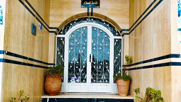 A hotel entrance with a white arched door, two large potted plants, and a sign above the door. - Apartment (fes)