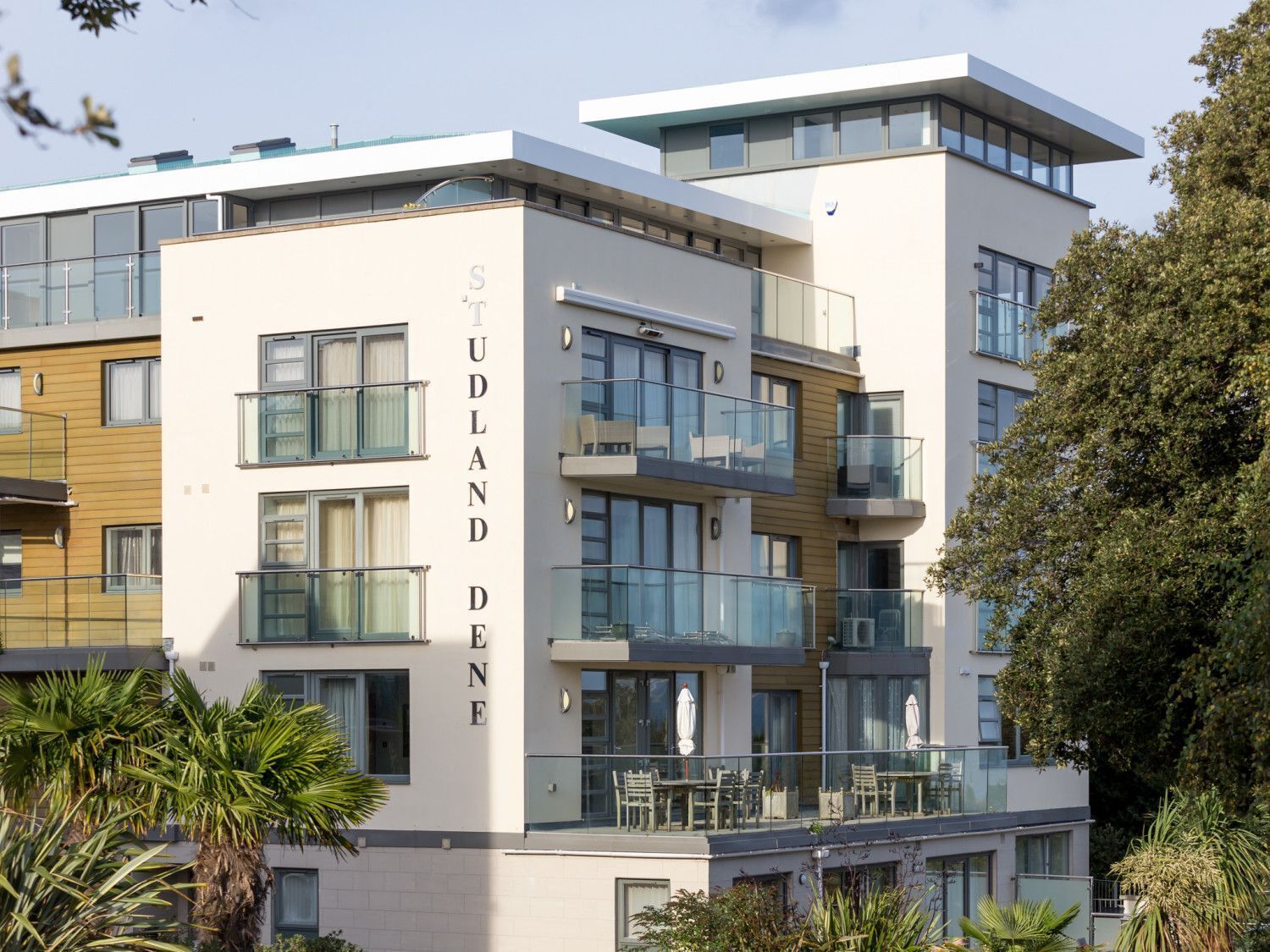 A modern multi-story building with balconies, large windows, and a sign reading 'STUDLAND DEN'.