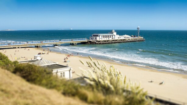 A beach with a pier extending into the ocean, a small building on the shore, and a clear blue sky.