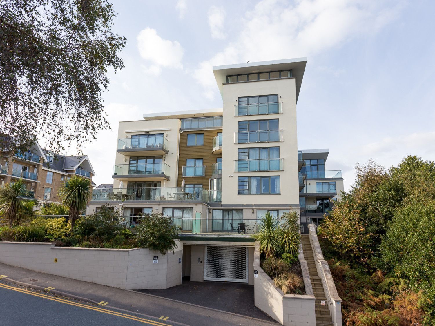 A modern multi-story residential building with balconies, a gated entrance, and surrounding greenery.