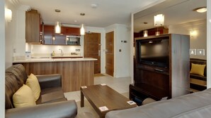 A modern living room with a sofa, a coffee table, and a TV. There is a kitchen area with wooden cabinets and a microwave.