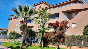 Un immeuble résidentiel à plusieurs étages, doté de balcons, d’un jardin agrémenté de palmiers et surplombant un ciel d’un bleu éclatant.