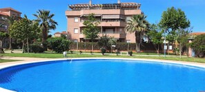 Uma piscina com superfície de água azul límpida, rodeada por um gramado bem cuidado e palmeiras. Ao fundo, um edifício de vários andares com varandas e telhado telhado.