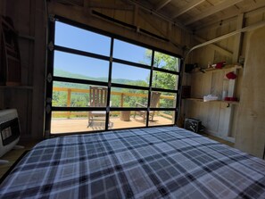 Relax in a cozy room with a large window overlooking a wooden deck and lush greenery, featuring a comfortable bed, a wooden chair, and a small table.