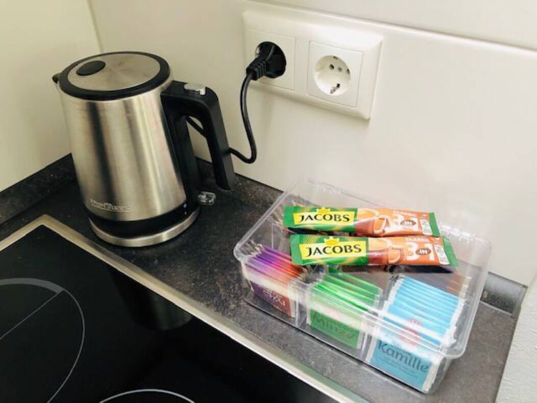 A kitchen counter with a kettle, a power outlet, and a plastic container with various food items.