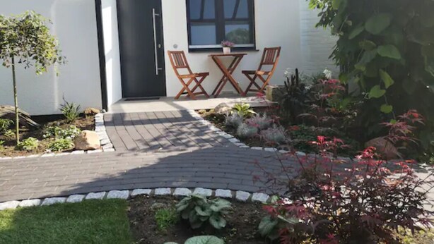 Un edificio moderno con una puerta marcada '54a', un ventanal pequeño y un camino pavimentado que lleva a una zona de estar con dos sillas y una mesa.