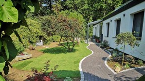 Un camino conduce a un edificio con un jardín que incluye césped, arbustos y un árbol pequeño.