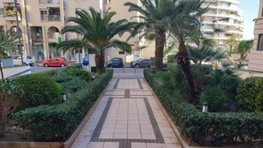 Un chemin pavé, bordé de haies soigneusement taillées et de palmiers, mène à un bâtiment doté de balcons.