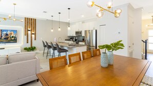 Eine moderne Küche mit Edelstahlgeräten, ein Essbereich mit Holztisch und -stühlen sowie ein Wohnzimmer mit Sofa und an der Wand montiertem Fernseher.