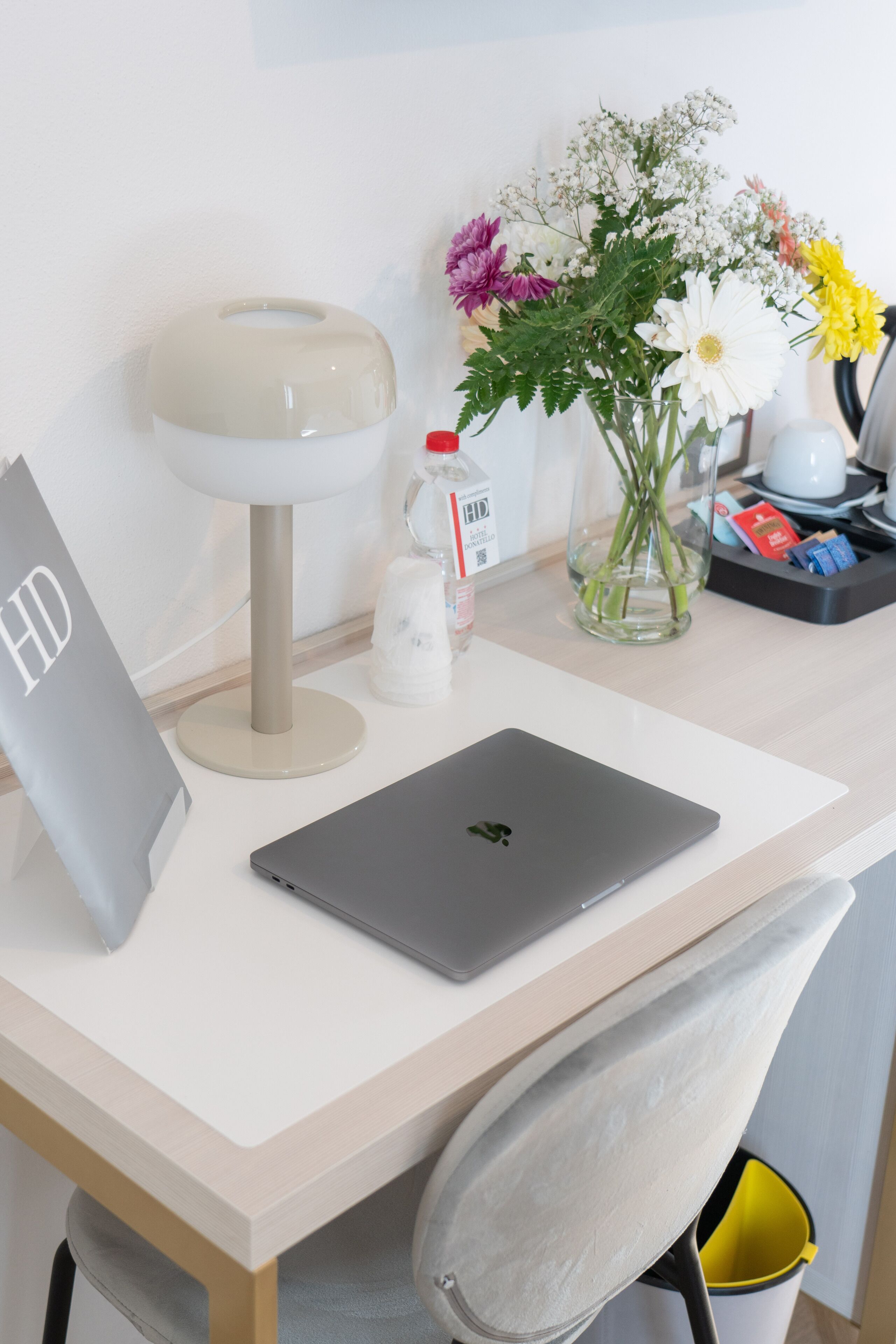 A desk with a laptop, a lamp, a vase with flowers, and a trash bin.