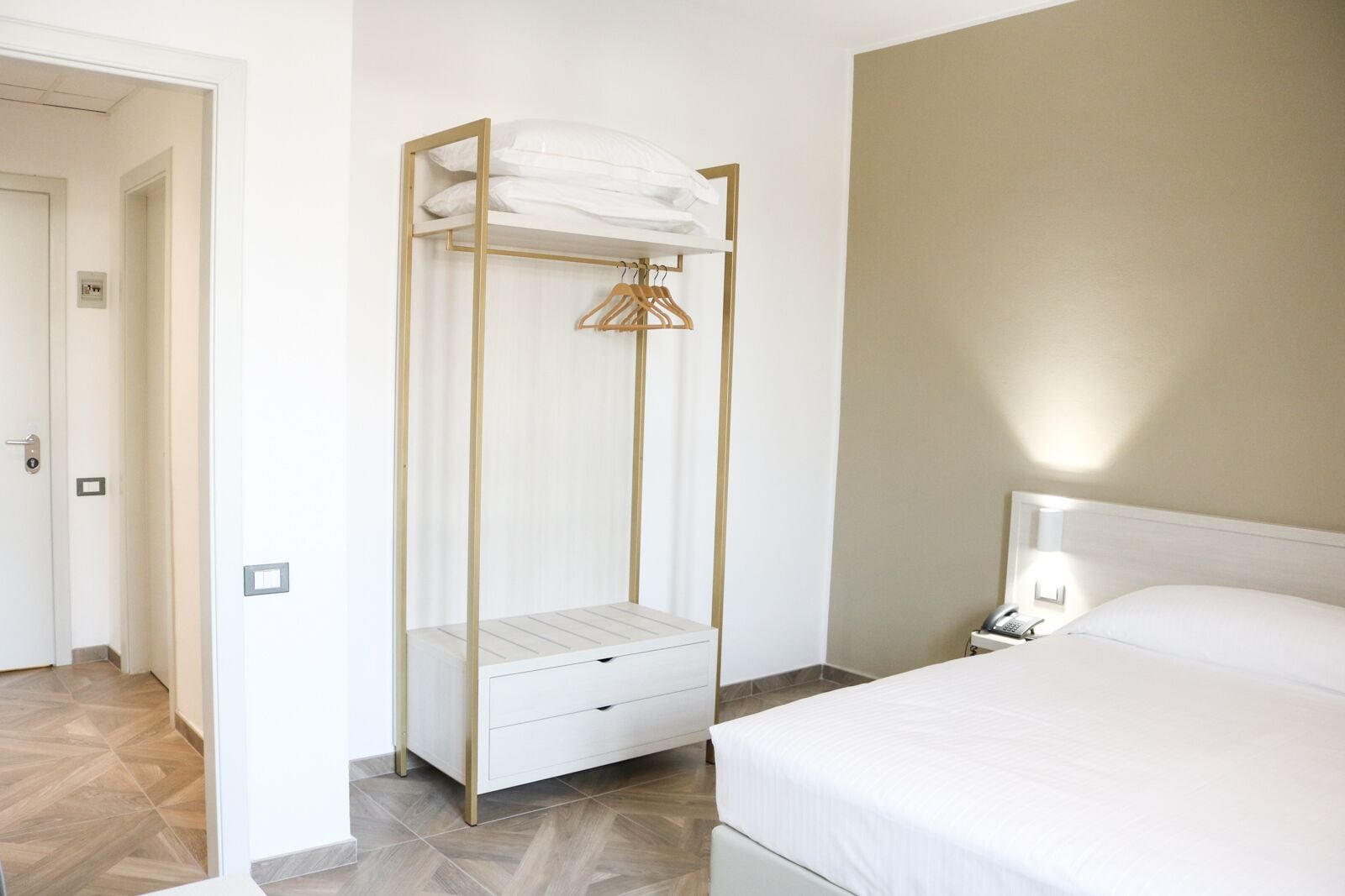 Habitación de hotel con cama, mesita de noche con lámpara, armario con perchero y puerta que da a otra habitación.