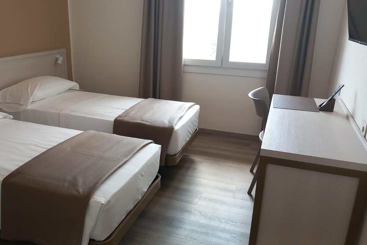 Een hotelkamer met twee eenpersoonsbedden, een klein bureau en een raam met gordijnen.