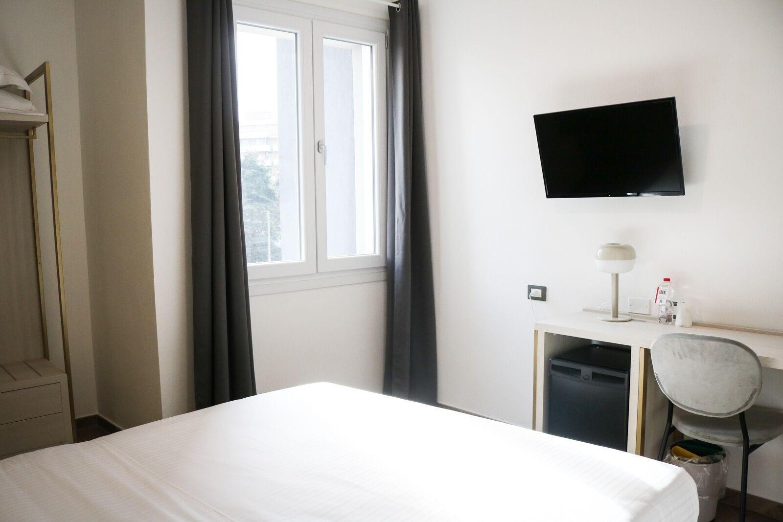 Habitación de hotel con cama, escritorio con silla, televisor montado en la pared y ventana con cortinas.