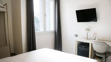 Habitación de hotel con cama, escritorio con silla, televisor montado en la pared y ventana con cortinas.
