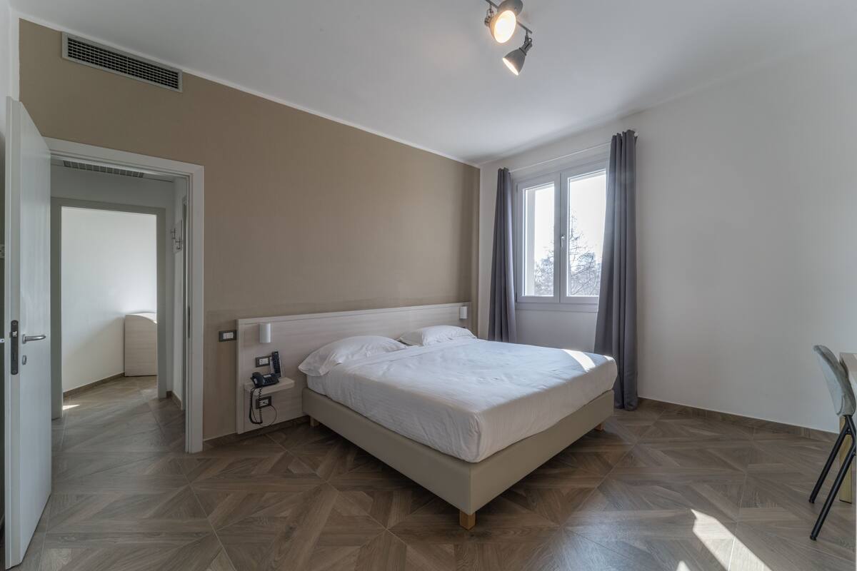 A bedroom with a bed, a bedside table with a lamp, a window with curtains, and a door leading to another room.