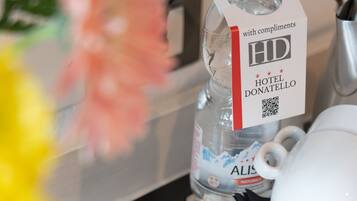 A hotel room tray with amenities including a water bottle, a white mug, a pair of black napkins, a red book titled 'Twinings', a blue packet labeled 'Of Light', and a white bag with a red logo.