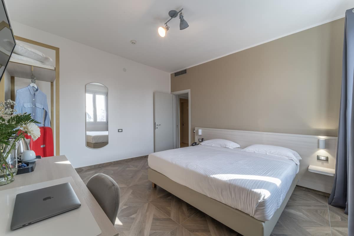 A hotel room with a bed, a desk with a laptop, a mirror, and a door leading to another room.
