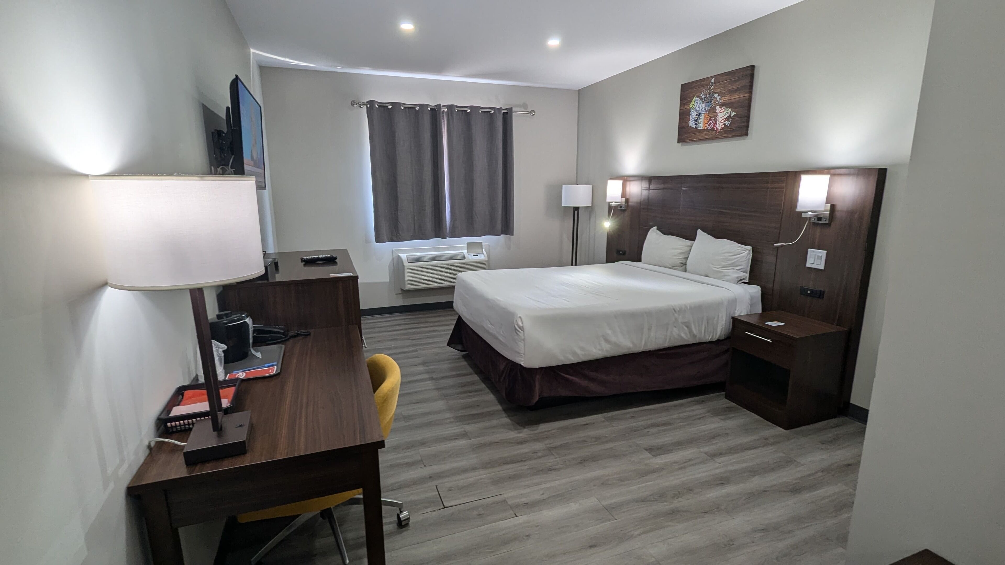 A hotel room with a bed, bedside tables, a desk with a lamp, a chair, a window with curtains, and artwork on the wall.
