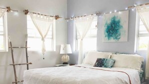 Une chambre à coucher avec un lit blanc, une table de chevet, une lampe et un tableau au mur.