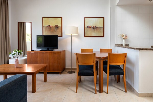 Une chambre d’hôtel avec une table à manger en bois, des chaises, un téléviseur, un meuble et des œuvres d’art accrochées aux murs.