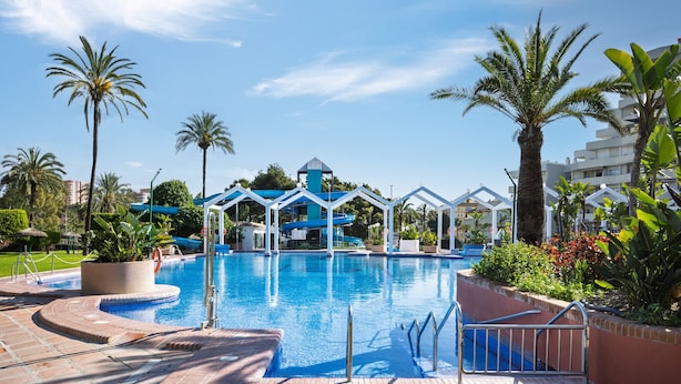 Una piscina con una estructura azul y blanca al fondo, rodeada de palmeras y un sendero de ladrillo.