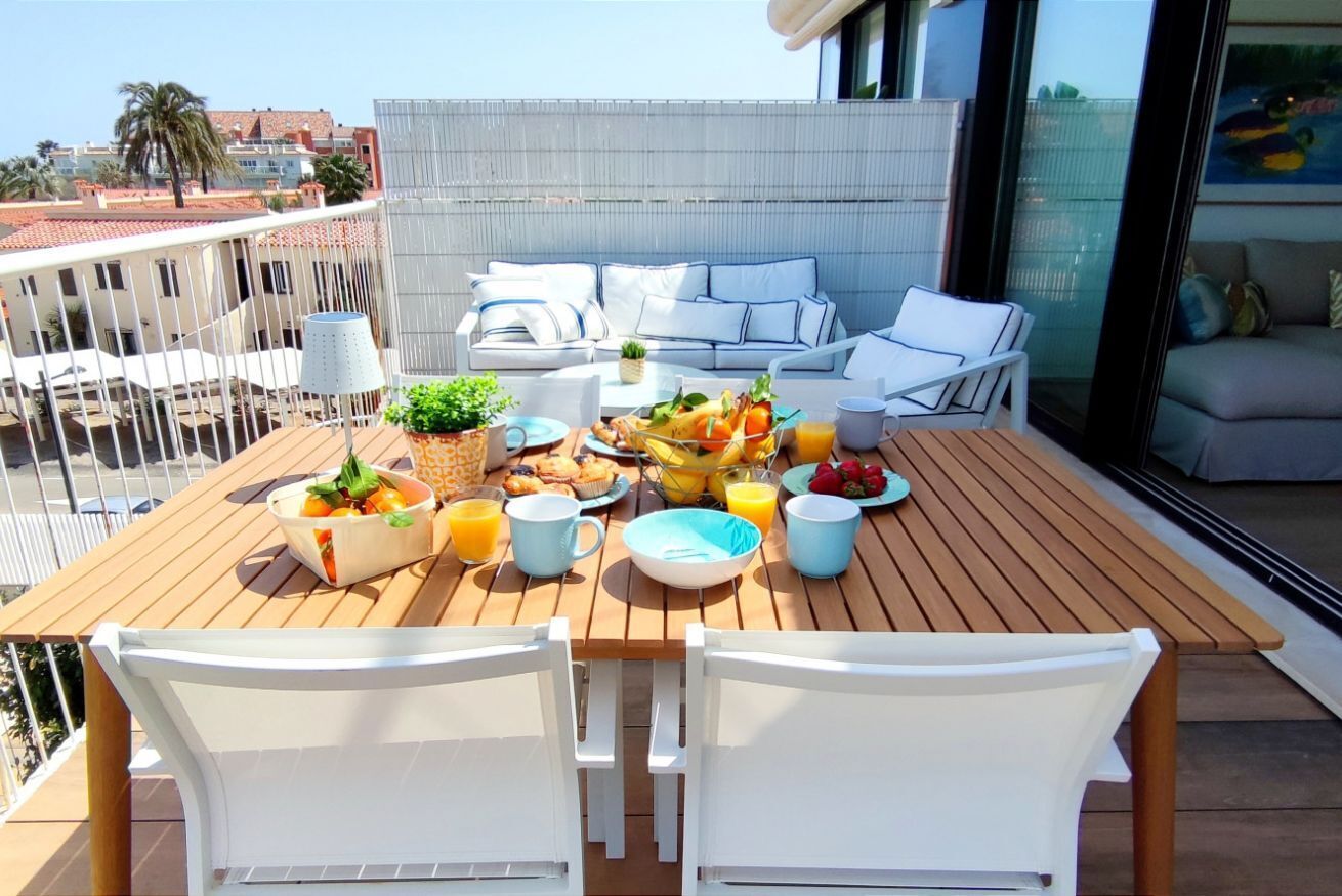 Une table en bois avec des aliments pour le petit-déjeuner, des chaises blanches et un canapé avec des coussins bleus, le tout sur un balcon extérieur.