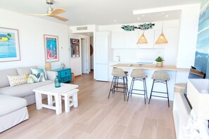 Una sala moderna con un sofá, una mesa de centro y taburetes de bar. Se ve una cocina con armarios blancos y luces colgantes.