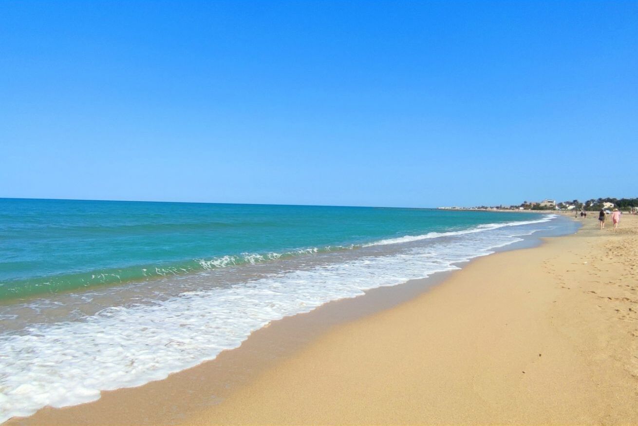 Une plage avec une eau d’un bleu limpide, un rivage sablonneux et quelques personnes qui se promènent.
