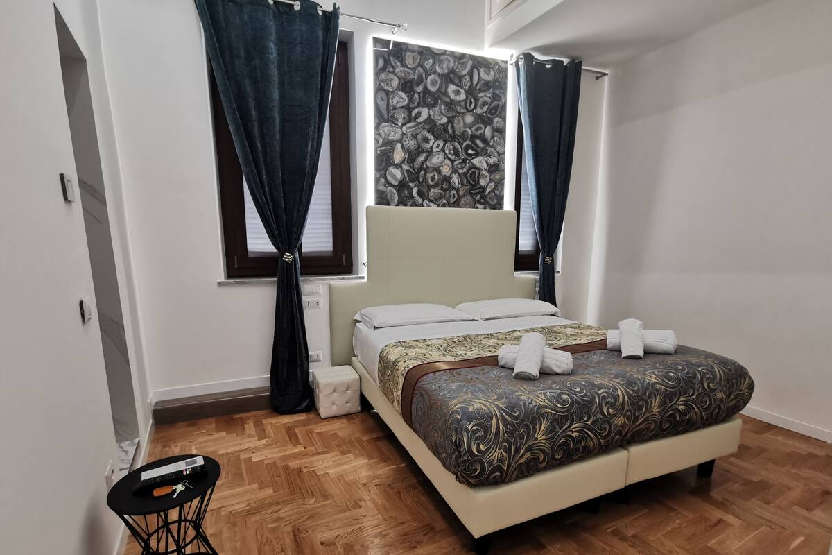 A bedroom with a bed, curtains, a window with a patterned curtain, a small table with a chair, and a wooden floor.