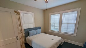 Una camera da letto con un letto, due finestre con tende, una porta e una ventola a soffitto.