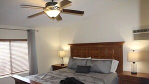 A bedroom with a wooden bed, two bedside tables with lamps, a ceiling fan, and a window with blinds.