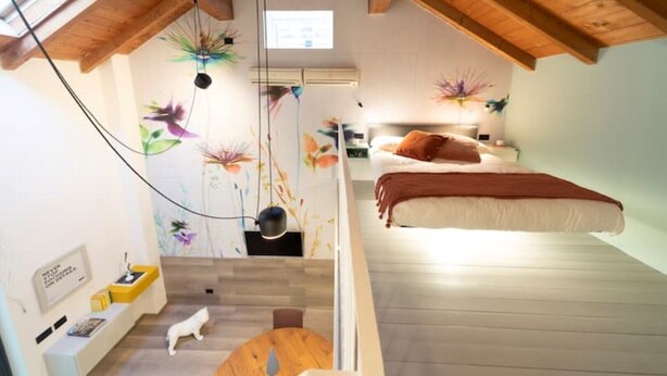 A loft-style bedroom with a sloped ceiling, a bed with a brown blanket, a small table with a white cat, and a wall with floral decorations.