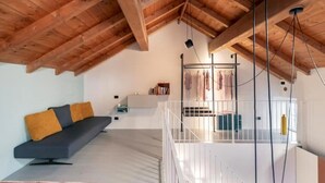 A modern loft apartment with exposed wooden beams, a grey sofa, a white staircase, and a balcony with clothes hanging.