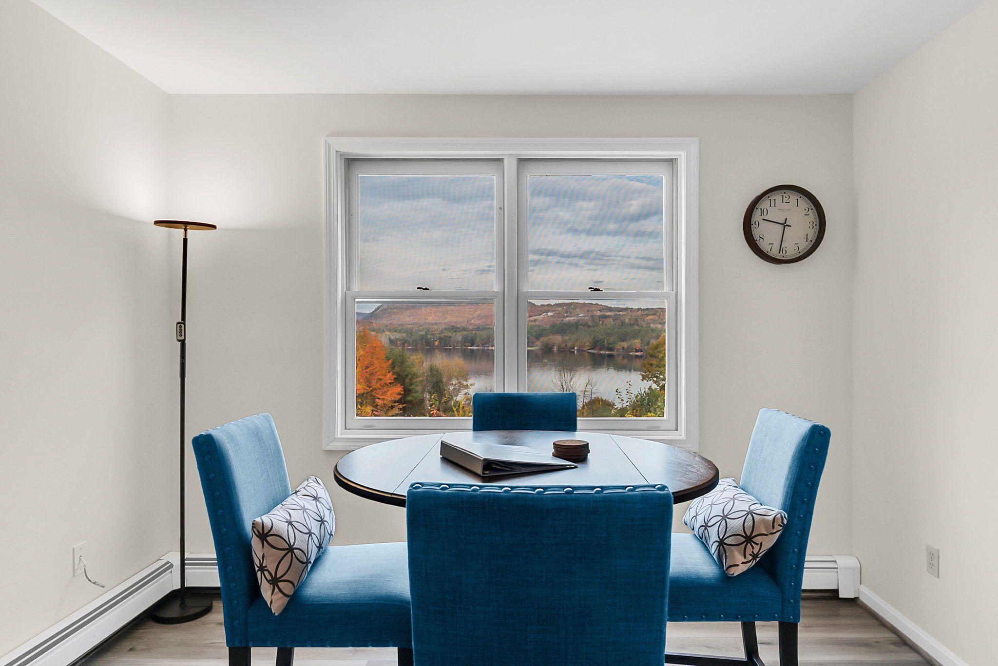 Un coin repas avec une table ronde et deux chaises bleues, une vue sur un lac et des collines, un lampadaire et une horloge au mur.