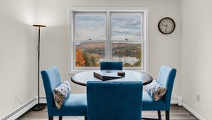 Een eethoek met een ronde tafel en twee blauwe stoelen, uitzicht op een meer en heuvels, een vloerlamp en een klok aan de muur.