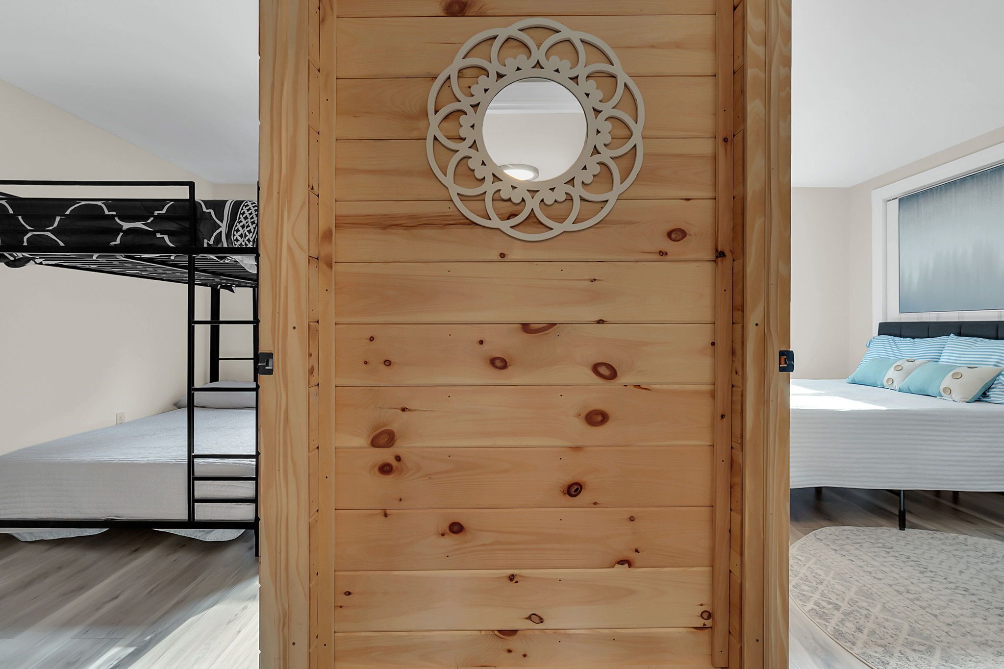 A room with a bunk bed, a wooden partition with a decorative mirror, and a bed with striped bedding.