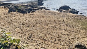 Een zandstrand met een houten pier die zich uitstrekt in kalm water, omringd door rotsen en schaarse begroeiing.
