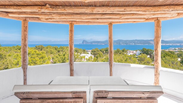 Una pérgola de madera con cojines blancos y vistas despejadas al mar y edificios lejanos.