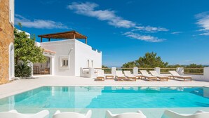 Un área de piscina con sillas reclinables blancas, un edificio de piedra y un cielo azul despejado.