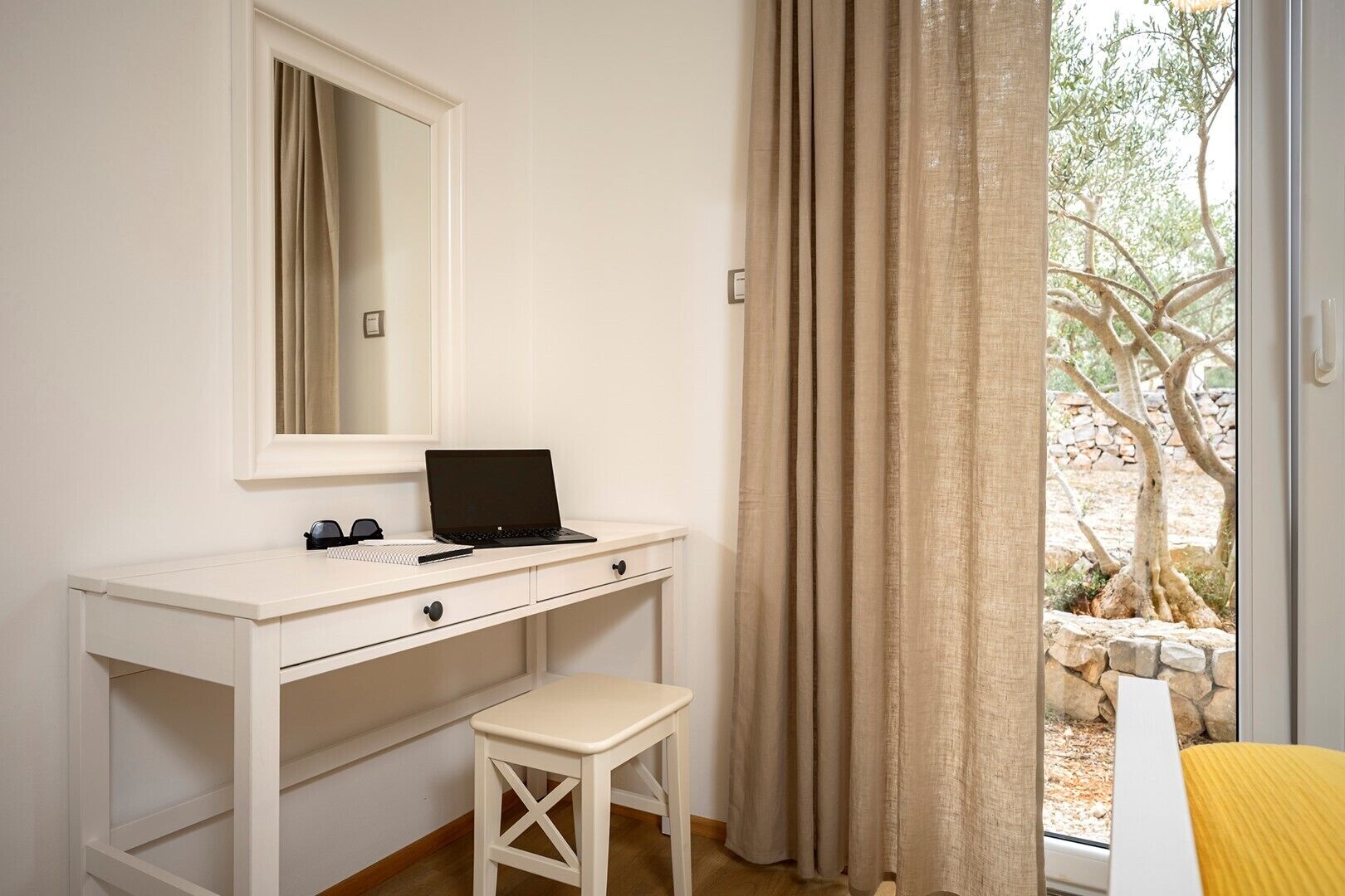 Une pièce avec un bureau blanc, un ordinateur portable, un petit tabouret, un miroir et une vue sur un espace extérieur avec des arbres et un mur en pierre.