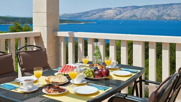 Une table dressée avec des aliments pour le petit-déjeuner, notamment du pain, des fruits et des boissons, offrant une vue panoramique sur la côte.