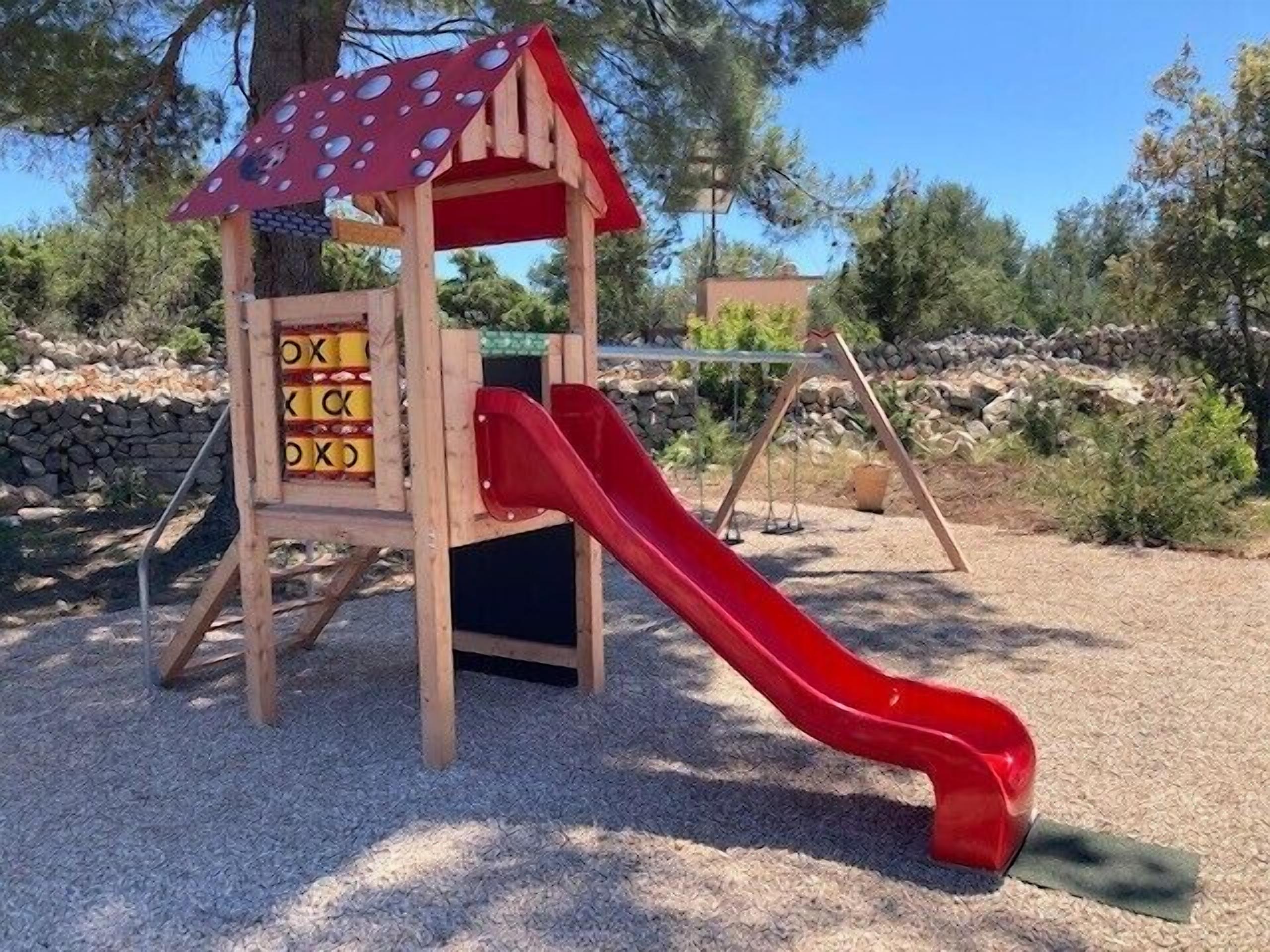 A wooden play structure with a slide, surrounded by gravel and trees.