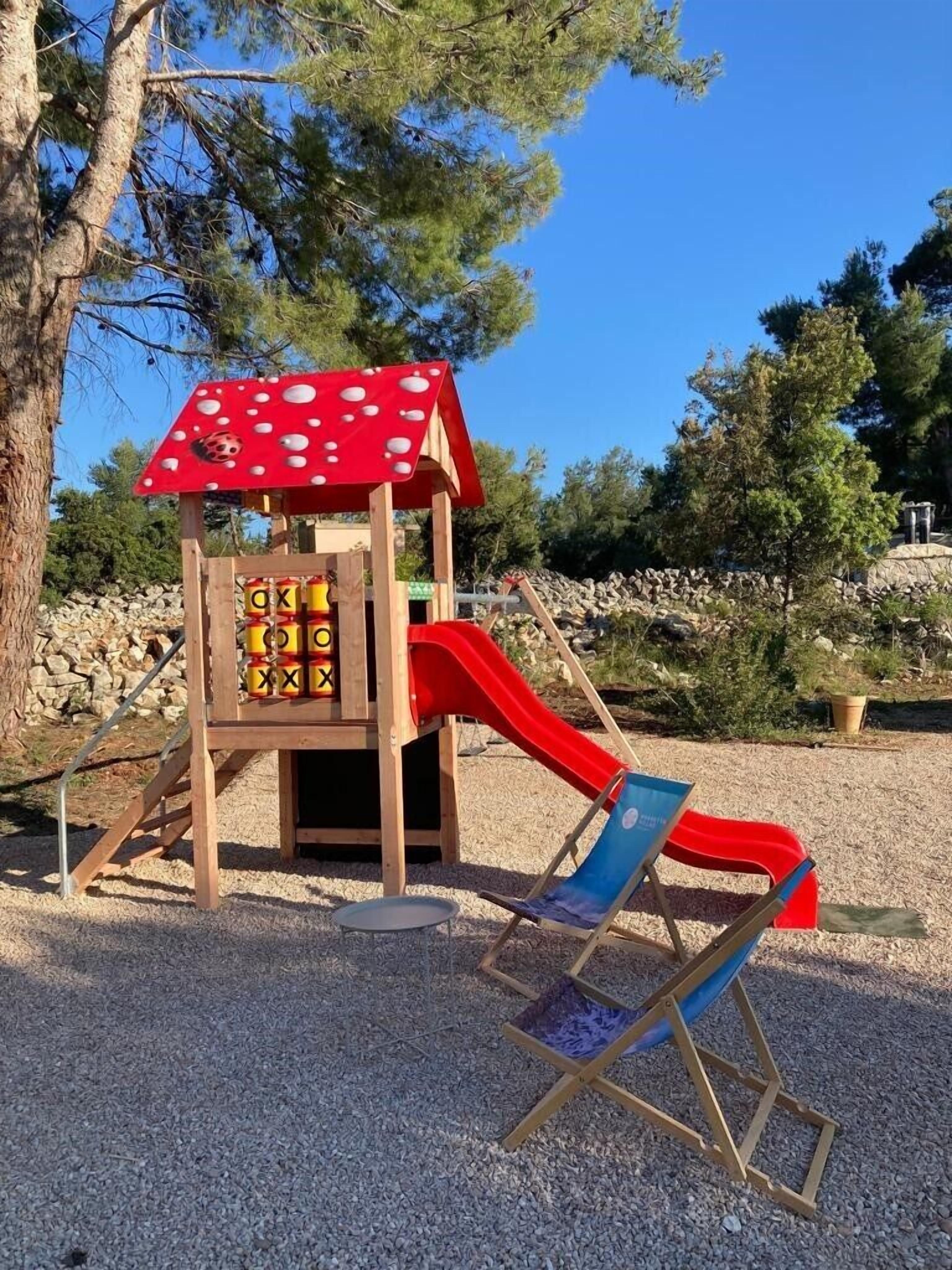 A children's play structure with a red slide and a climbing wall, surrounded by trees and gravel.