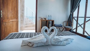 A bedroom with a bed, a heart-shaped towel arrangement, a wooden wardrobe, a small table with bottles, a chair, and a view of the sea through large windows. - ZAN Tropical Jambiani Beach (Jambiani)