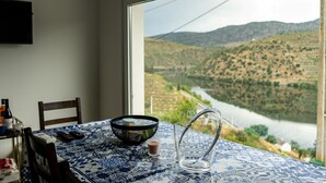 Une table à manger recouverte d’une nappe à motifs bleus et blancs, un pichet en verre transparent, un saladier noir en treillis et un petit verre contenant une boisson rouge. Une fenêtre offre une vue sur une rivière et un paysage vallonné.