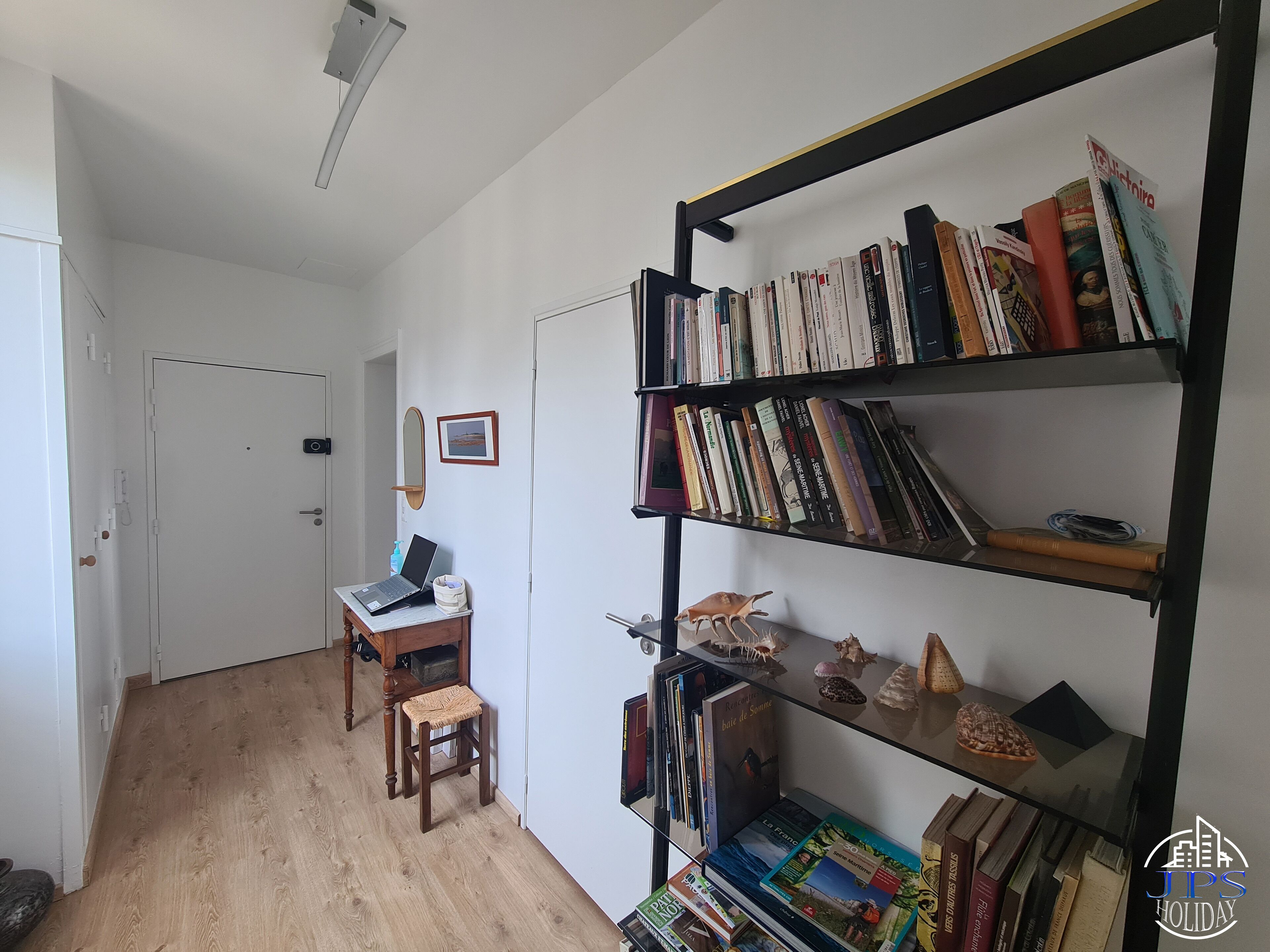A room with a wooden floor, a desk with a laptop, a bookshelf filled with books, and a small stool.