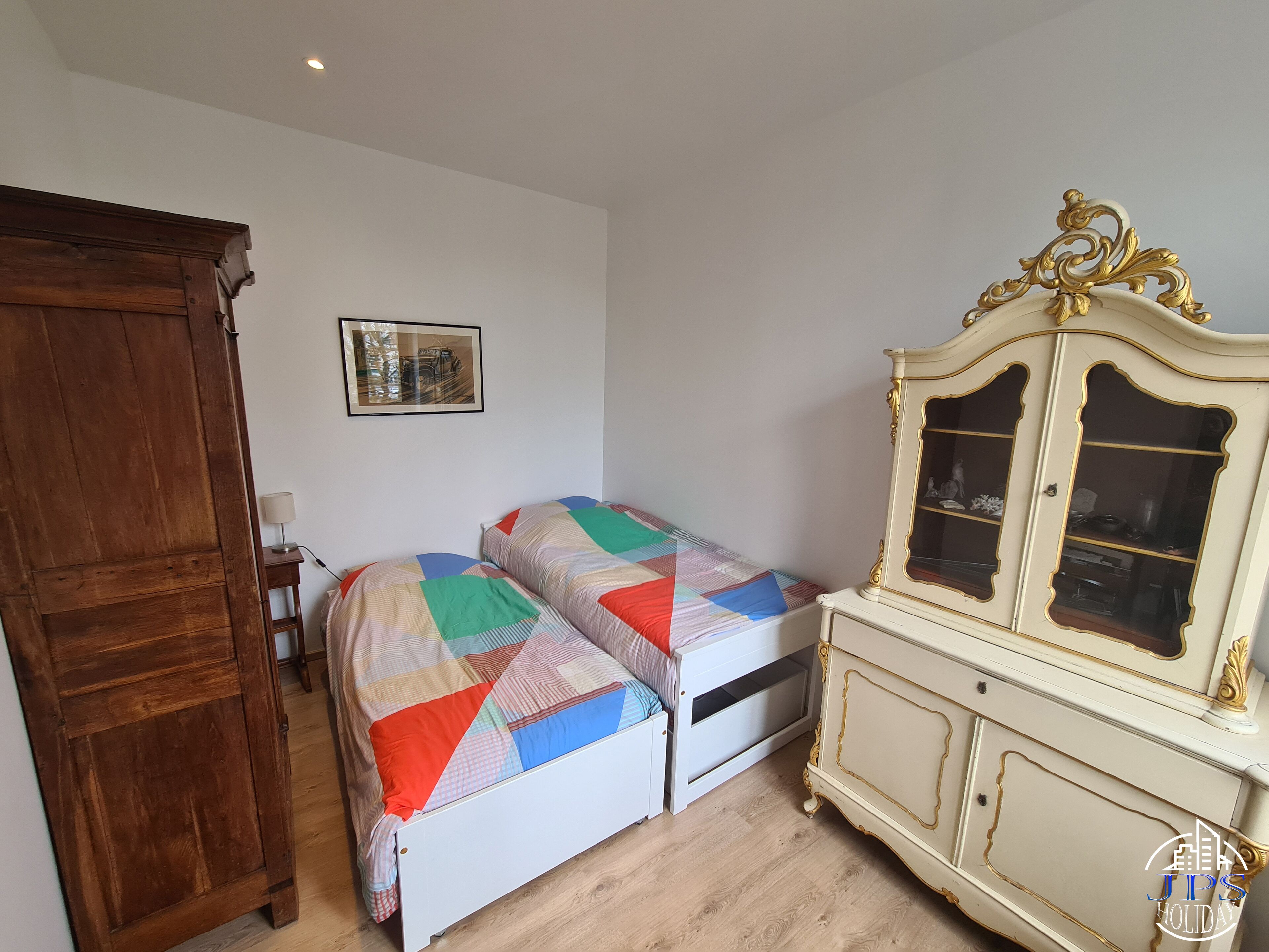 A bedroom with two beds, a wooden wardrobe, a white cabinet with glass doors, and a framed picture on the wall.