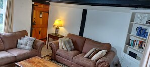 A cozy living room with two brown leather sofas, a wooden coffee table, a small side table with a lamp, and a bookshelf filled with books and decorative items.