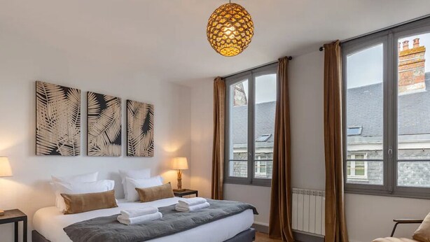 Ett hotellrum med en säng, två sängbord, ett stort fönster med gardiner och en dekorativ taklampa.